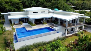 A modern two-story house with a large swimming pool, surrounded by lush greenery and a well-maintained garden.