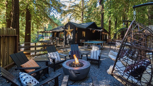 An outdoor seating area with a fire pit, surrounded by wooden furniture and a rustic cabin in the background. - Casa Boheme-Stylish, Relaxing, Close to Downtown! (Guerneville)