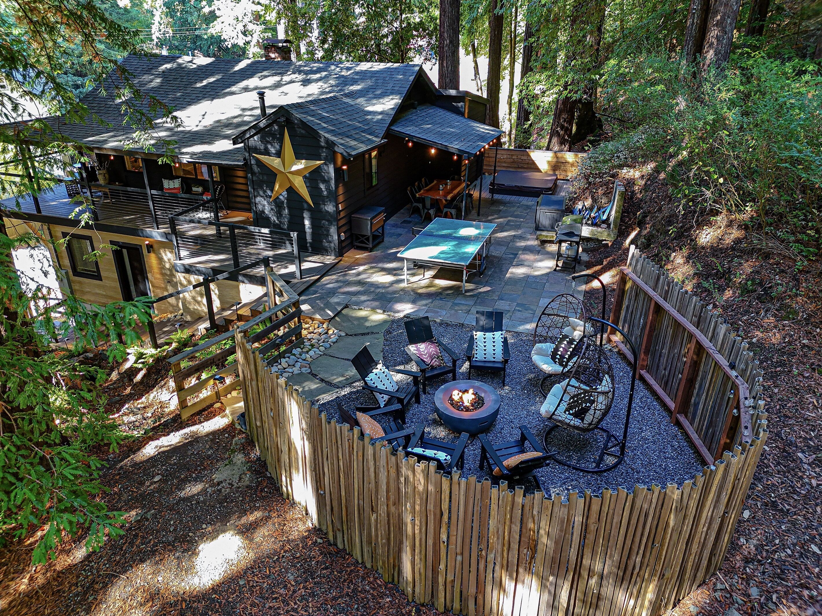 Eine Holzhütte mit sternförmigem Dach, Sitzbereich im Freien mit Stühlen und Feuerstelle, umgeben von einem Holzzaun und Bäumen.