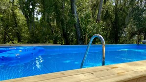 Ein Schwimmbecken mit Holzterrasse und Metallleiter, umgeben von Bäumen.