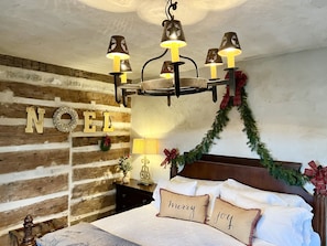 Un dormitorio acogedor con una pared de troncos de madera, una lámpara de araña, una cama con almohadas que dicen 'Merry' y 'Joy', y una decoración navideña que dice 'Noel'.