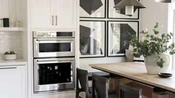 A modern kitchen with white cabinets, stainless steel appliances, a wooden dining table, and a potted plant.