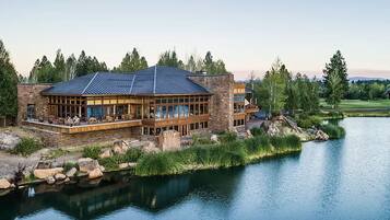 A large, multi-story lodge with a stone facade and extensive glass windows, situated by a calm lake with a golf course in the background.