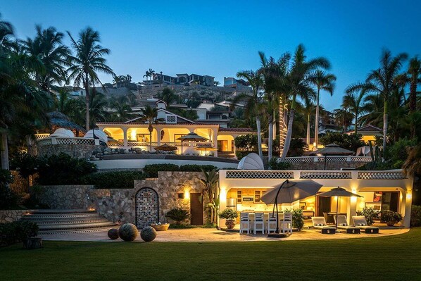A multi-story resort with outdoor seating, palm trees, and a stone wall entrance. - Casa Edwards: 8BR Beachfront Palmilla Estate with Gym, Pool & Event Space (San Jose del Cabo)