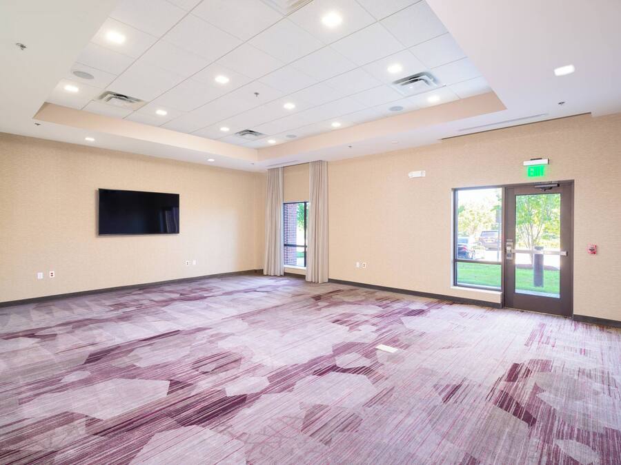 A large, empty conference room with a patterned carpet, beige walls, a flat-screen TV mounted on the wall, and a door leading outside.