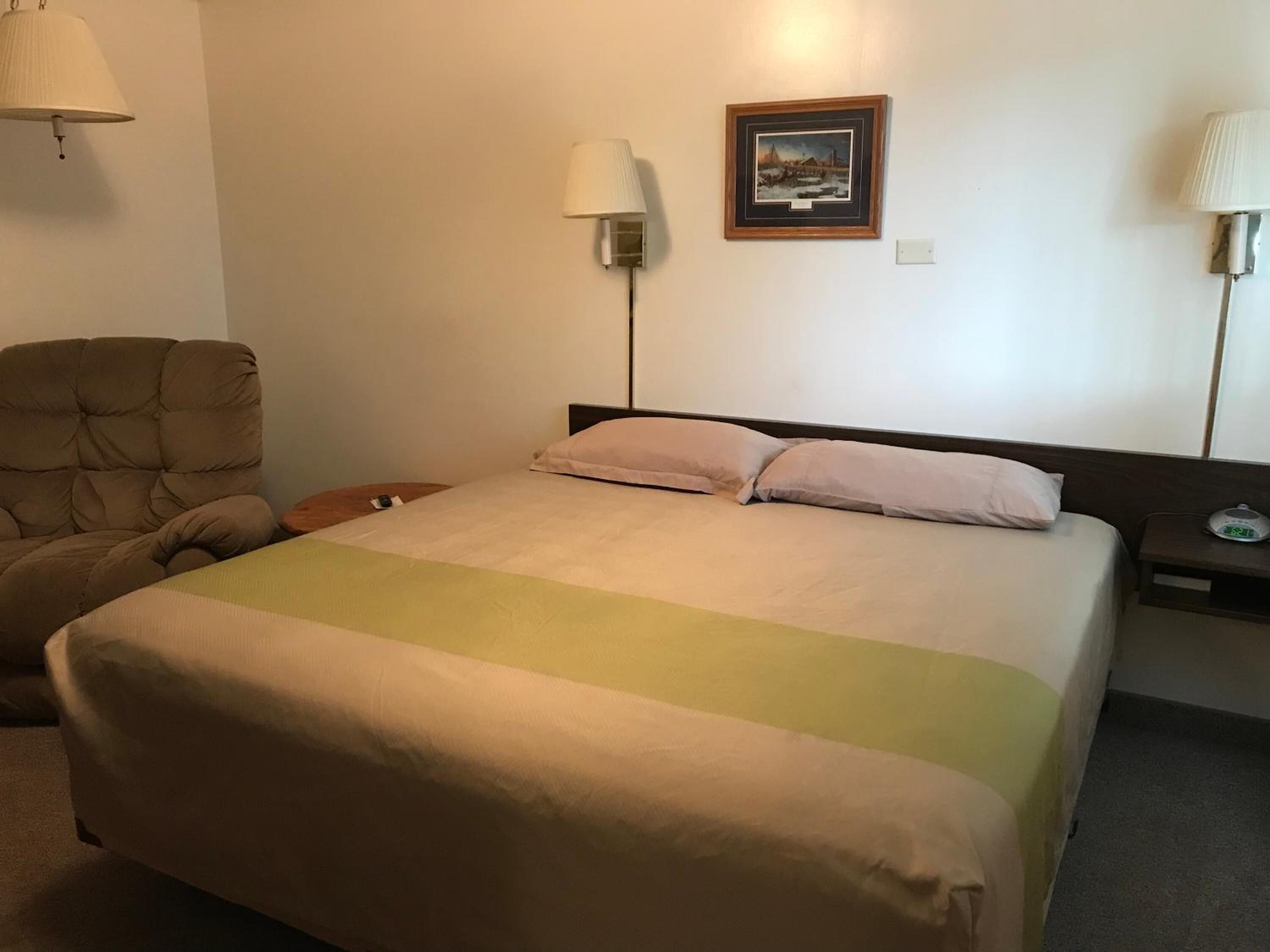 A hotel room with a bed, a chair, two bedside lamps, a nightstand, and a framed picture on the wall.