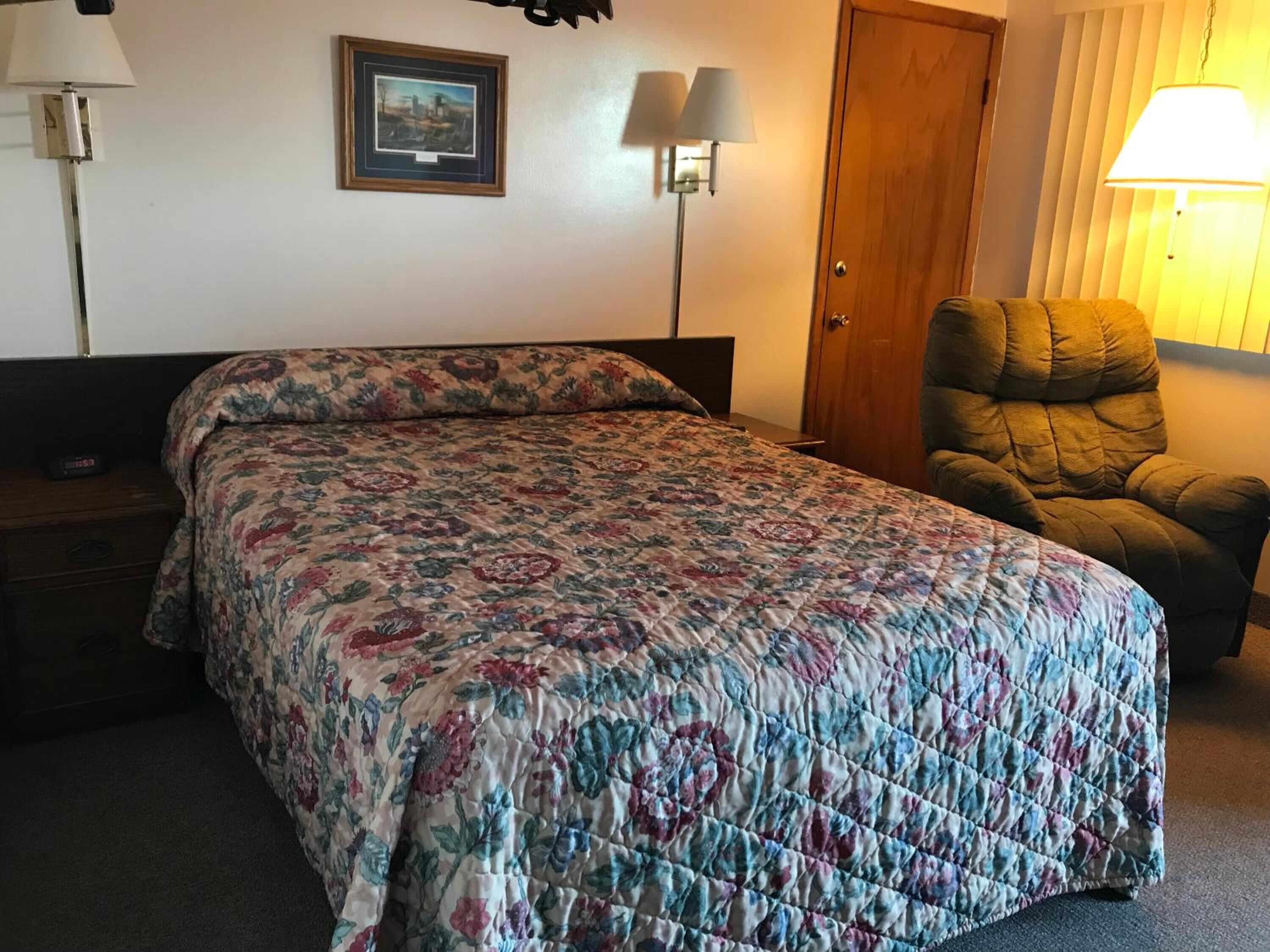 A hotel room with a bed, a chair, a nightstand, and two wall-mounted lamps.