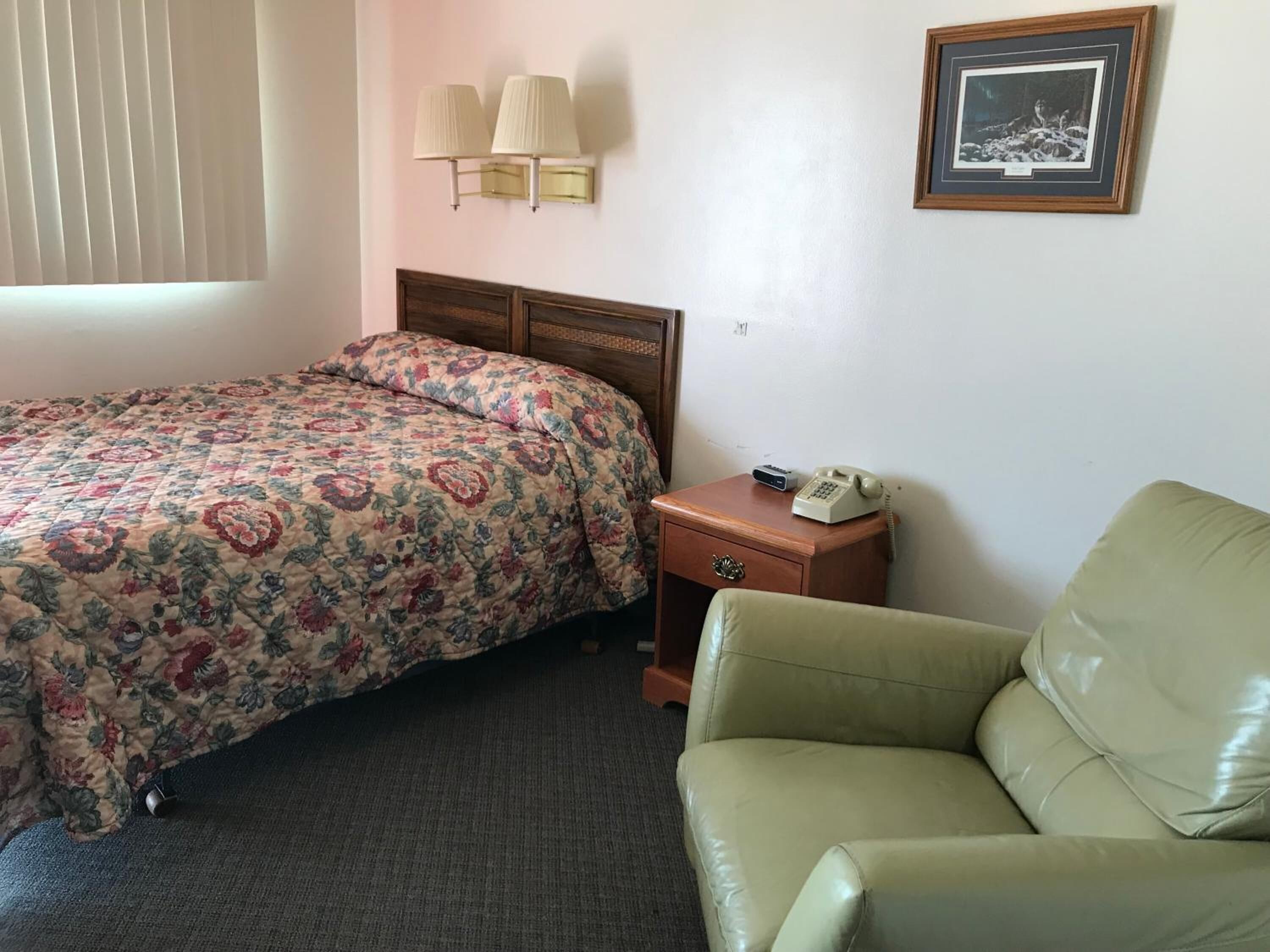 A hotel room with a floral bedspread, a wooden nightstand with a telephone, and a green armchair.