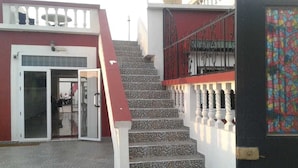 Una escalera conduce a un edificio con fachada roja y blanca, puerta de entrada de vidrio y un balcón con barandilla metálica.