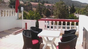Una terraza con muebles de mimbre y una mesa blanca, con vistas a un paisaje montañoso.