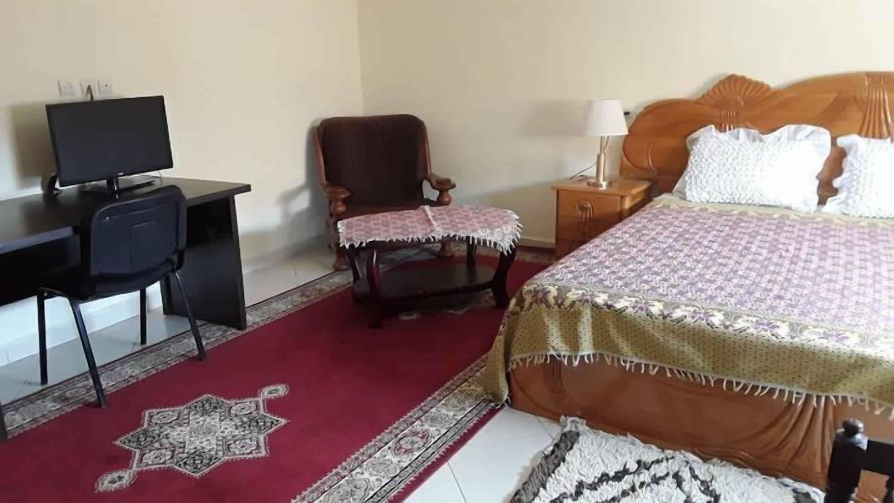 A hotel room with a bed, a desk with a computer, a chair, a nightstand, and a lamp.
