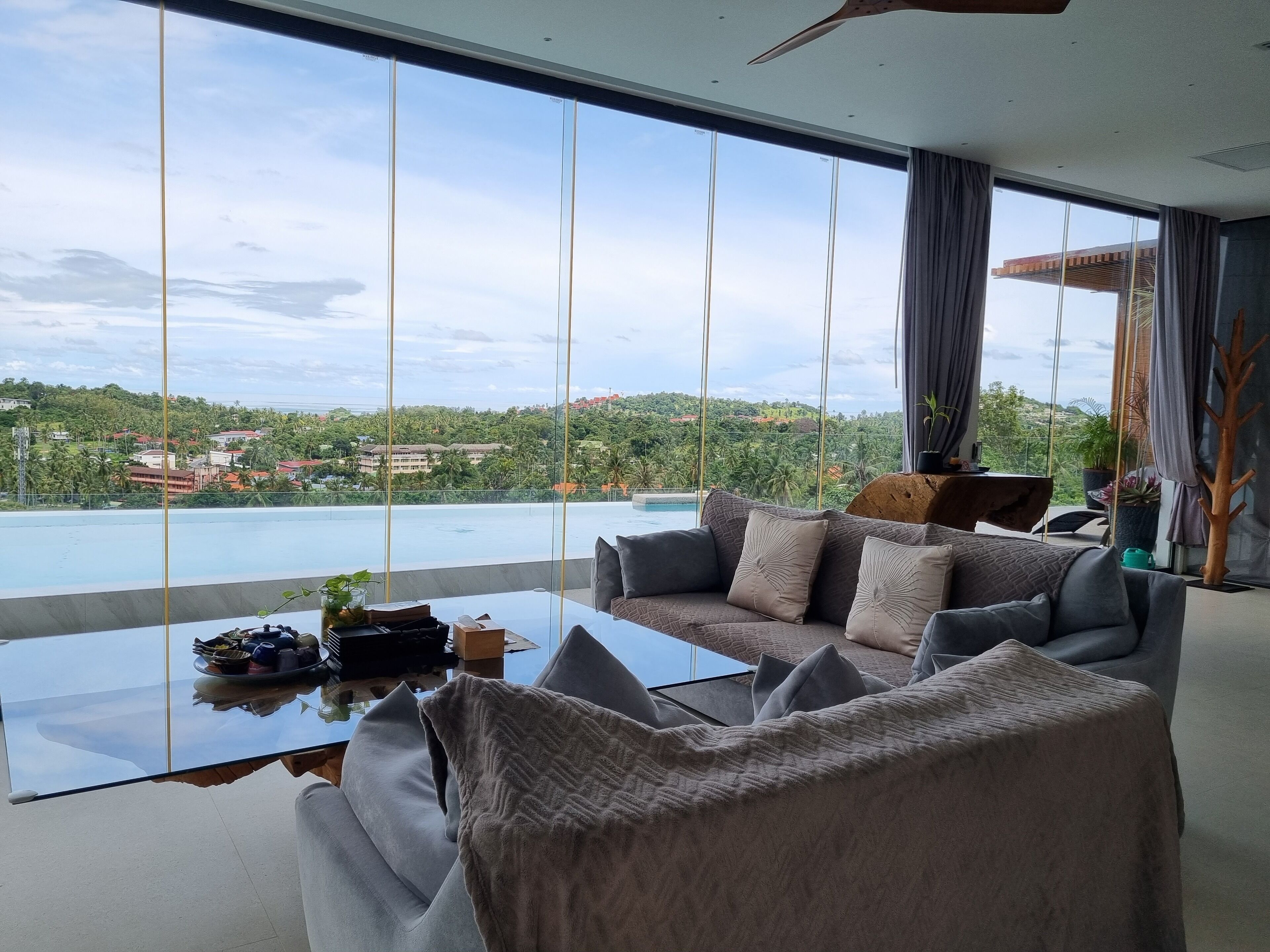 Sala de estar moderna com uma mesa de centro de vidro grande, sofá cinza e vista para uma piscina e áreas verdes.