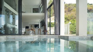 Ein moderner, überdachter Pool mit Glasboden, eine Küchenzeile mit Barhocker und große Fenster mit Blick auf die umliegende Natur.