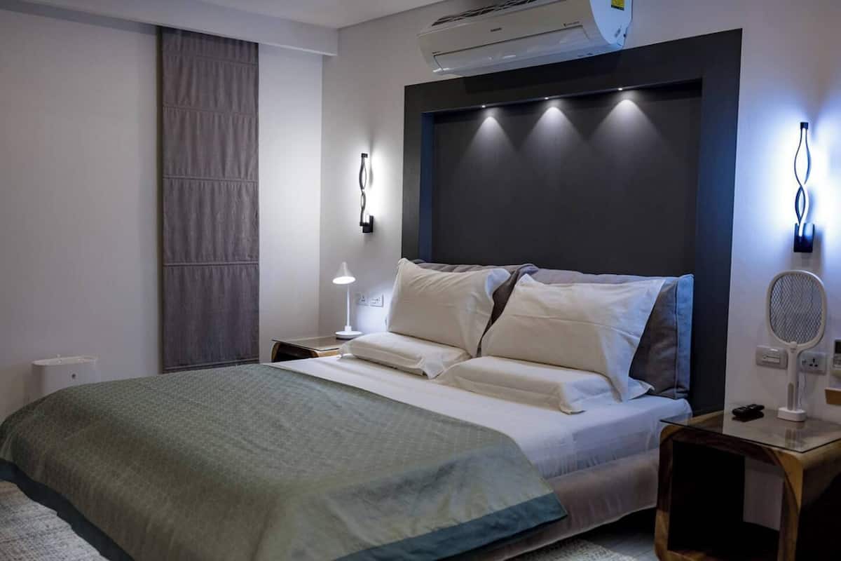 A modern hotel room with a large bed, a bedside table with a lamp, an air conditioning unit, and a wall-mounted light fixture.