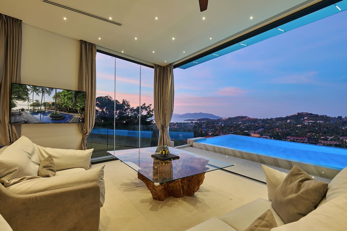 A modern living room with a glass coffee table, beige sofas, and a large TV screen.