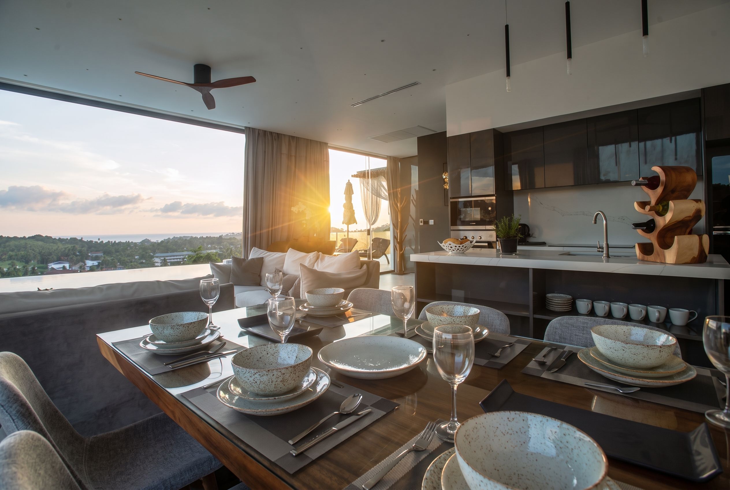A modern dining area with a table set for four, a kitchen with dark cabinetry, and a view of the landscape.