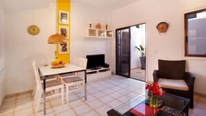 Un espacio habitable compacto con una mesa de comedor, sillas, un televisor, un sofá, una pequeña estantería y una planta en maceta.