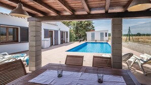 Un espace extérieur abrité comprenant une piscine, des meubles en bois et une pergola agrémentée de guirlandes lumineuses.
