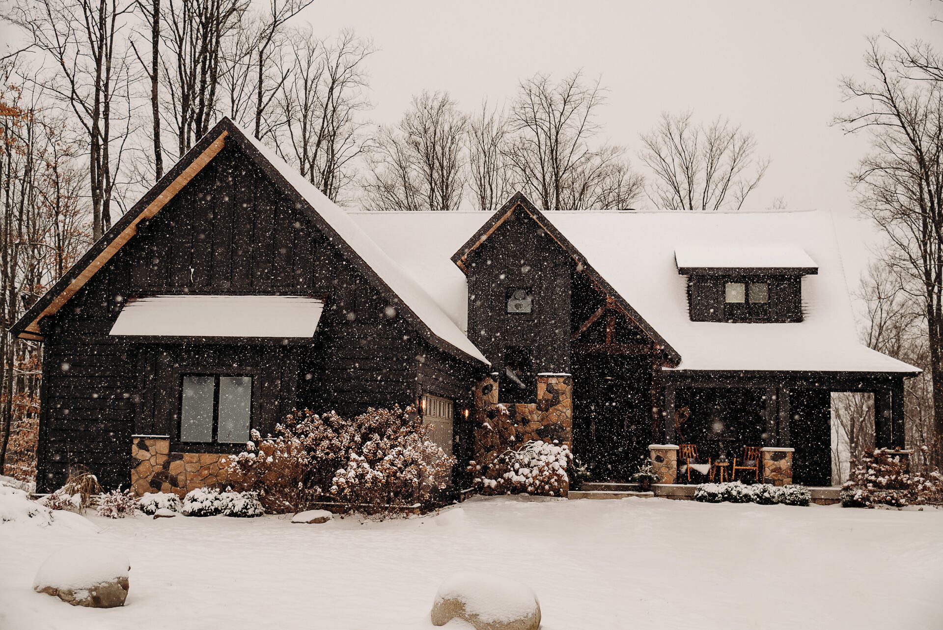 A snow-covered house with a stone foundation, a large chimney, and a snow-covered roof.