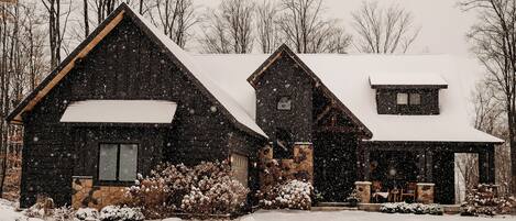 A snow-covered house with a stone foundation, a large chimney, and a snow-covered roof.