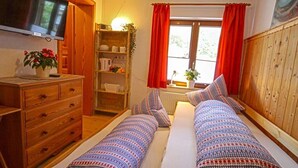 A cozy room with two beds, a wooden dresser, a TV, and a window with red curtains. - Fewo Bergsteigerkammerl, 1 Person, Terrasse, 12 qm (Schönau am Königssee)