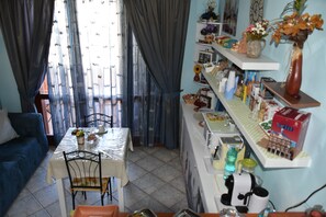 A dining area with a table set for two, a blue sofa, and a kitchen area with shelves and appliances. - Il Girasole (Assemini)