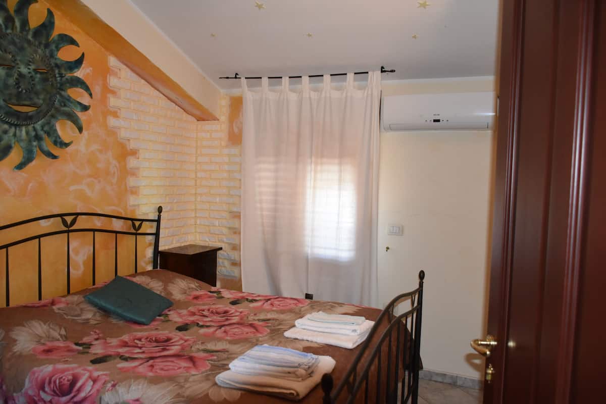 Un dormitorio con cama, mesita de noche, decoración en la pared, ventana con cortinas y una puerta.