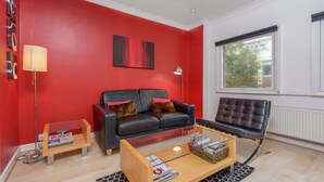 A modern living room with a red accent wall, a leather sofa, a leather armchair, a glass coffee table, and a floor lamp. - Super Central London - sleeps 4 (London)