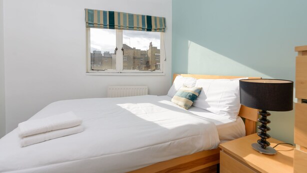 A neatly made bed with white linens, a striped window valance, and a bedside lamp on a wooden nightstand. - Super Central London - sleeps 4 (London)