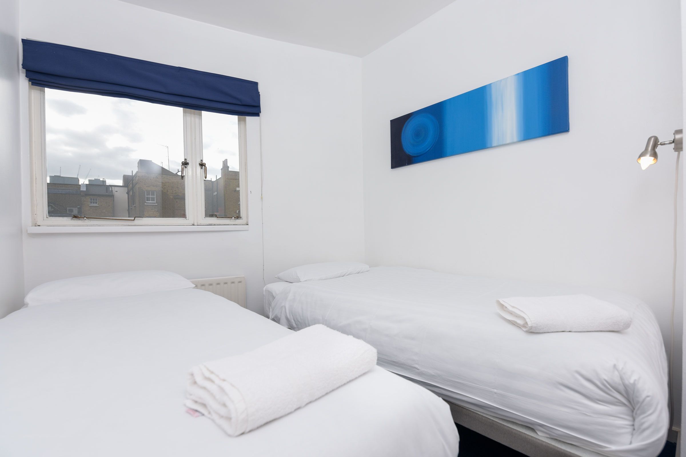 Ett hotellrum med två sängar, ett fönster med en blå persienn, en abstrakt tavla som hänger på väggen och en sänglampa.