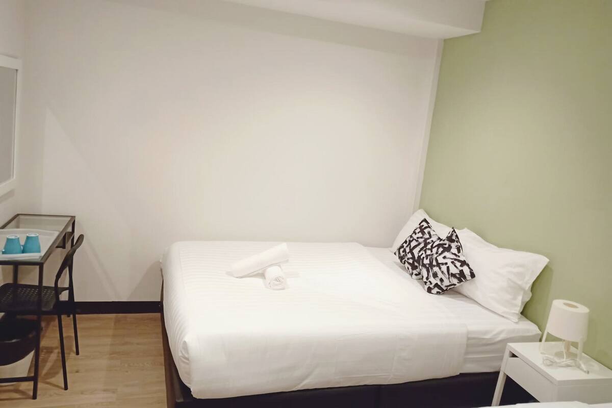A neatly made bed with white linens, a black and white patterned pillow, and a rolled white towel. There is a small table with a chair and a blue cup on it. A white nightstand with a lamp is visible.