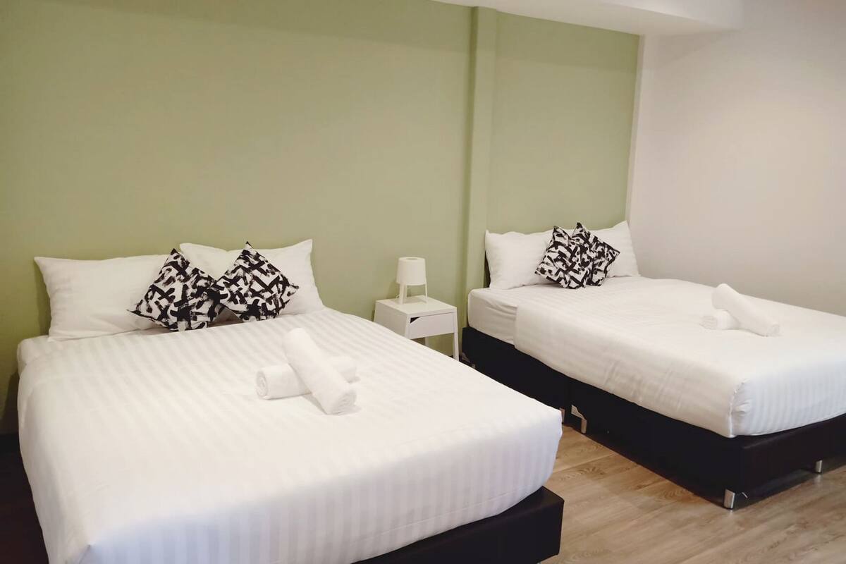 A hotel room with two beds, each with white bedding and black patterned pillows. There is a small bedside table with a lamp on one side and a white box on the other. The walls are painted in a light green color.