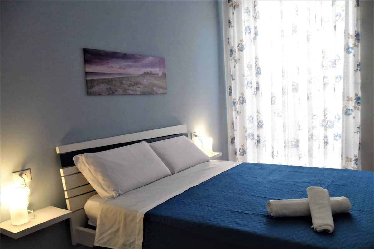 A neatly made bed with white and blue bedding, a white pillow, and a rolled towel on the bed. There is a nightstand with a lamp and a power outlet on the left. A painting is hung on the wall above the bed.