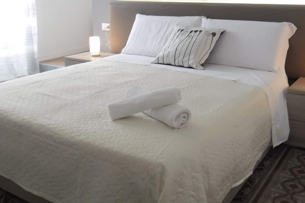 A neatly made bed with white linens and a rolled towel, a bedside lamp, and a patterned rug.
