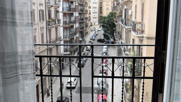 A balcony with a view of a narrow street lined with multi-story buildings, featuring balconies and parked cars. - Le vie del Centro (Palermo)