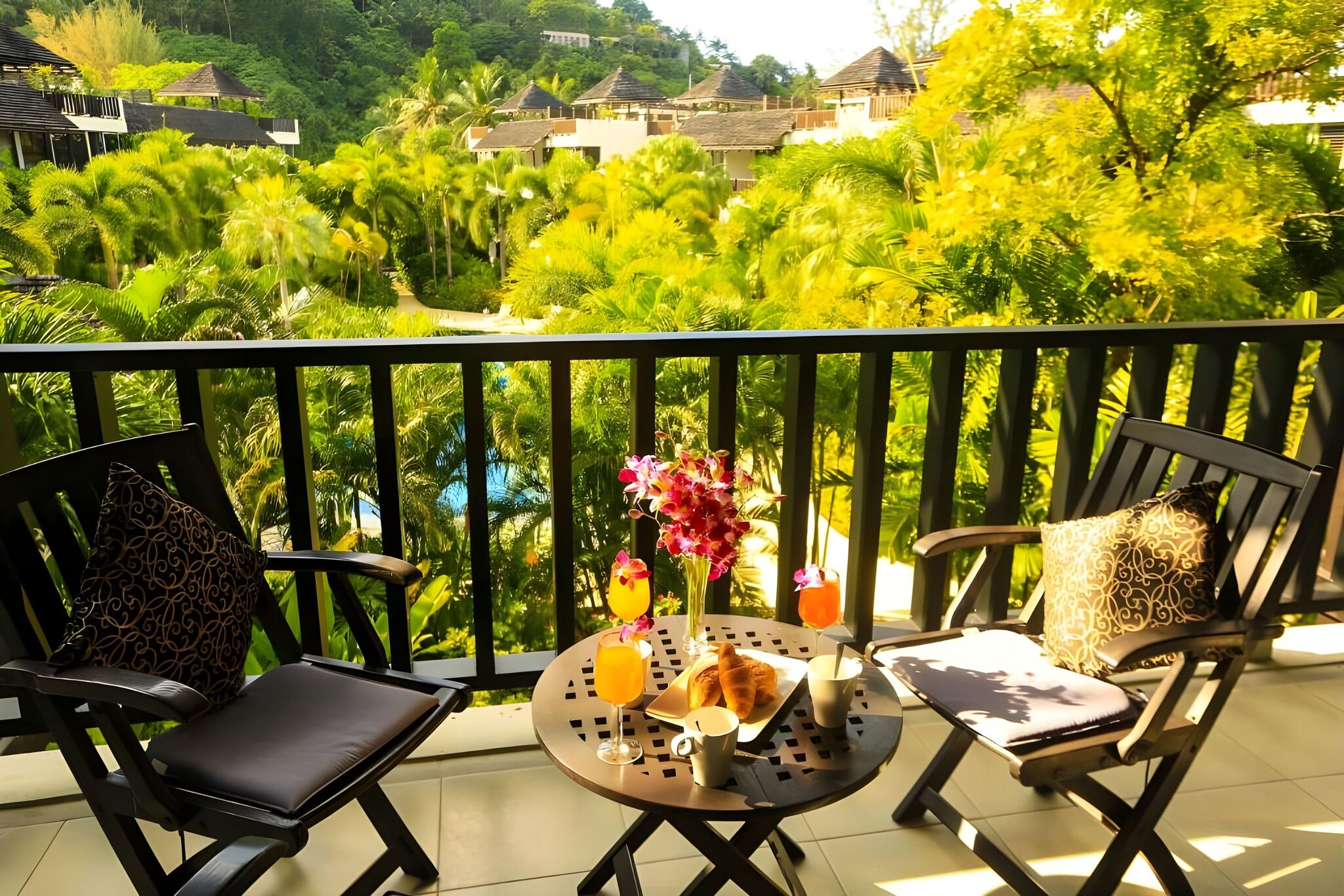 A balcony with two chairs, a table with a drink, a flower vase, and a croissant.