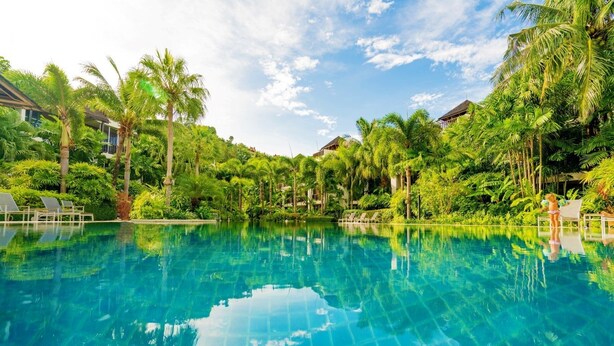 A clear blue pool surrounded by lush greenery and palm trees, with lounge chairs and a building in the background. - Bangtao Beach Garden By RESAVA (Choeng Thale)