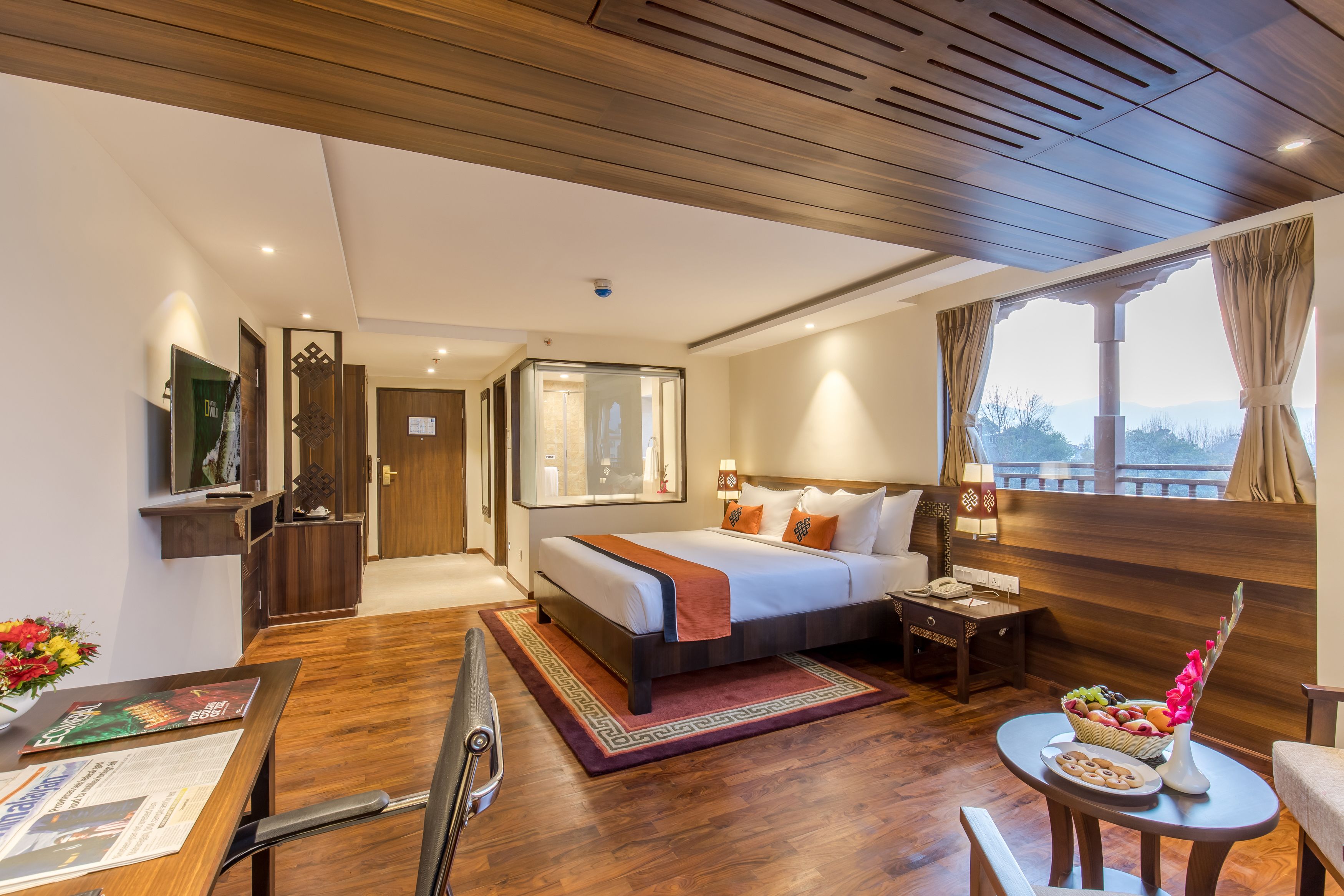 Habitación de hotel con una cama grande, un escritorio con silla, una mesita con un florero y frutas, y una ventana con cortinas.