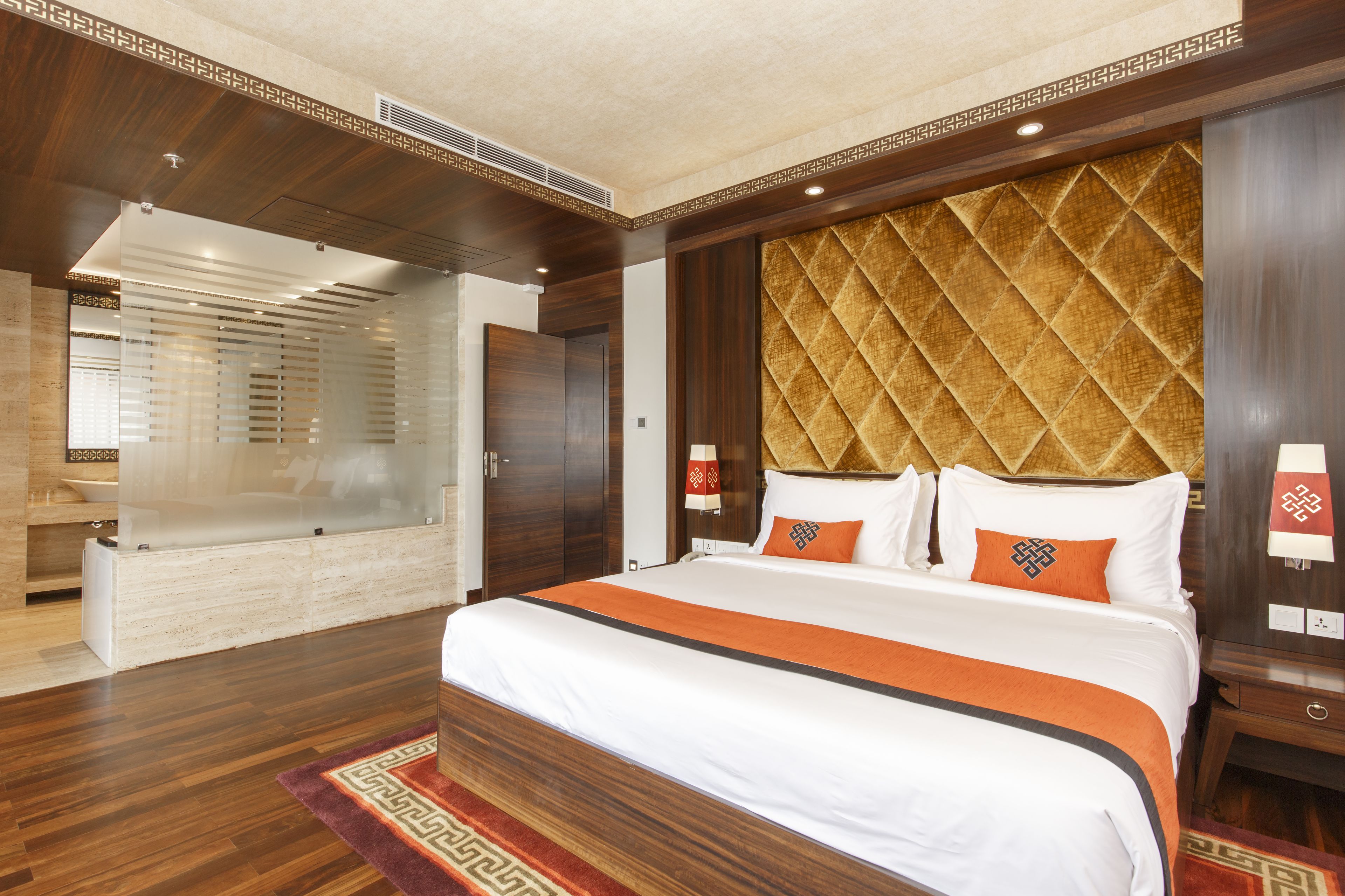 Een hotelkamer met een groot bed, een houten hoofdbord, een badkamer met een glazen douchewand en een vloerkleed met een rood-wit patroon.