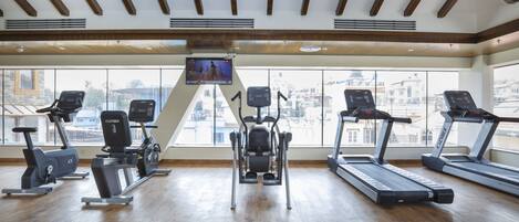 Une salle de sport équipée de divers appareils d’exercice, dotée de grandes fenêtres et offrant une vue sur la ville.