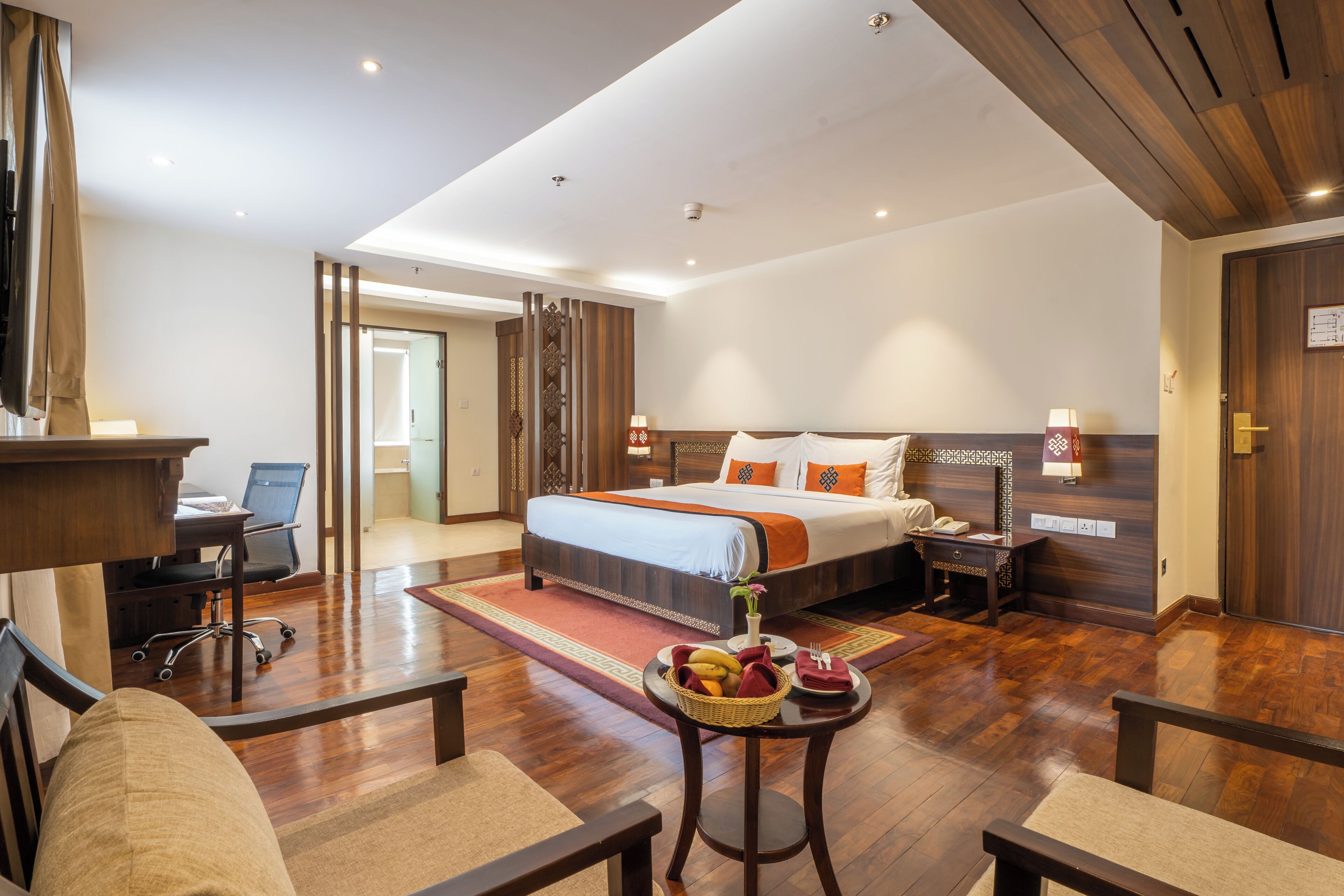 A hotel room with a bed, desk, chair, and a small table with a fruit basket.