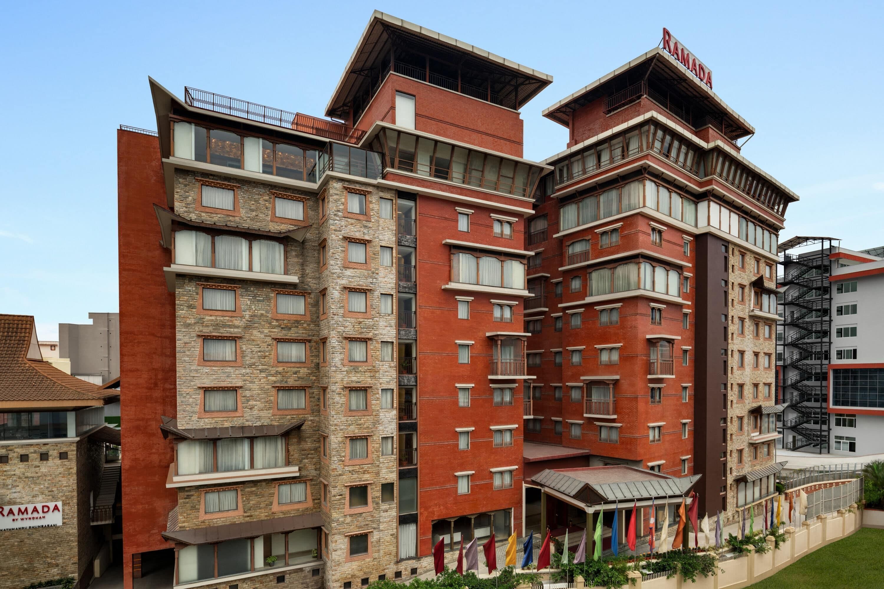 A multi-story hotel with a modern design, featuring a mix of brick and stone facades, multiple balconies, and a prominent entrance with flags.