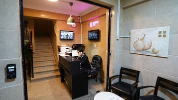 A hotel reception area with a desk, chairs, a staircase, and a wall-mounted TV.