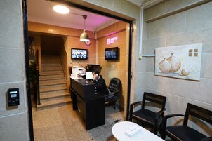Zona de recepción de un hotel con mostrador, sillas, una escalera y un televisor en la pared.