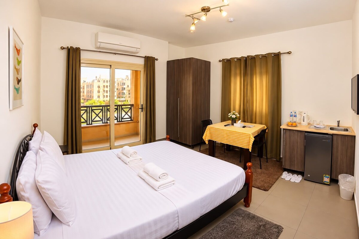 A hotel room with a bed, a small table with a yellow tablecloth, a kitchenette, and a balcony with curtains.