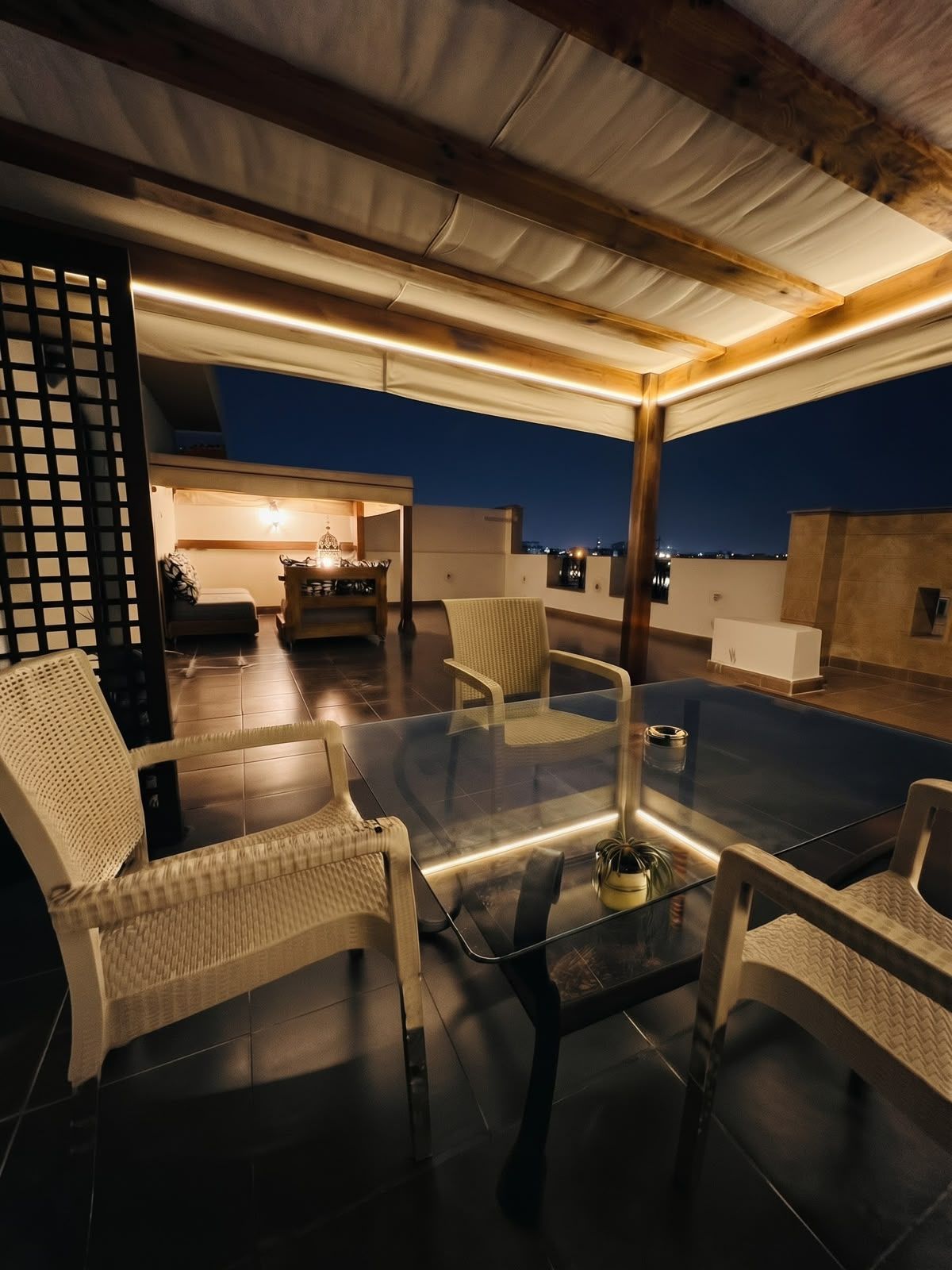 Una terraza en la azotea con una mesa de vidrio y sillas de mimbre, vista de la ciudad al anochecer, y una zona de estar acogedora con una lámpara.