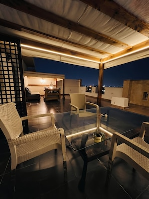 A rooftop terrace with a glass table and wicker chairs, a view of the city at dusk, and a cozy seating area with a lamp. - Sakan 275 (New Cairo)