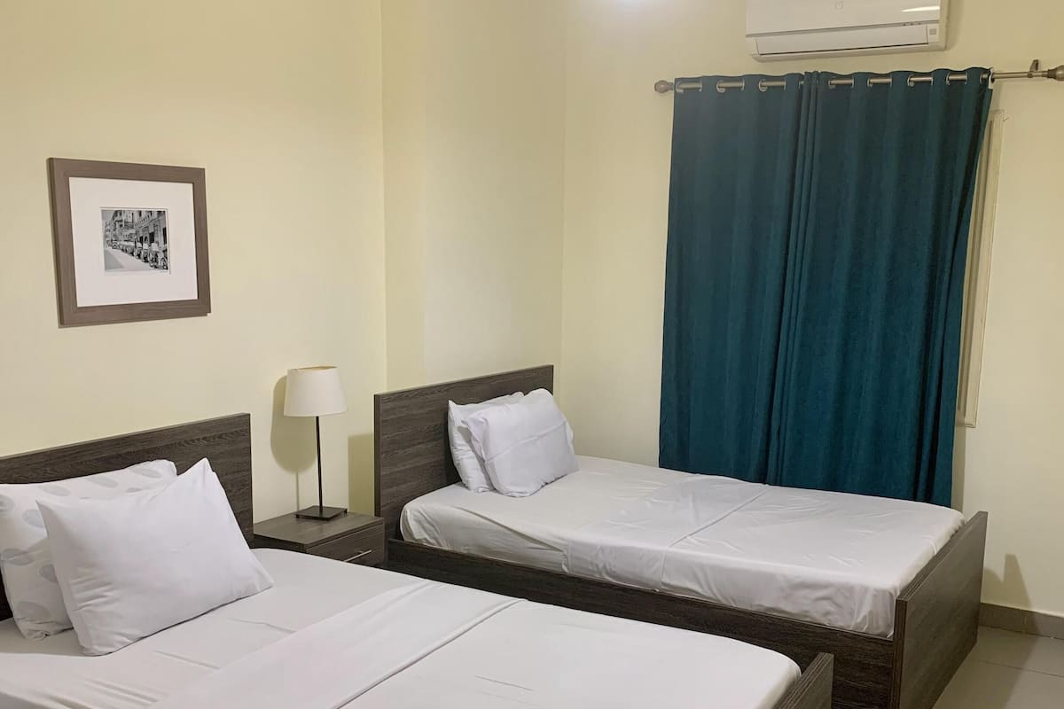 A hotel room with two single beds, a nightstand with a lamp, a framed picture on the wall, an air conditioning unit, and a curtain.