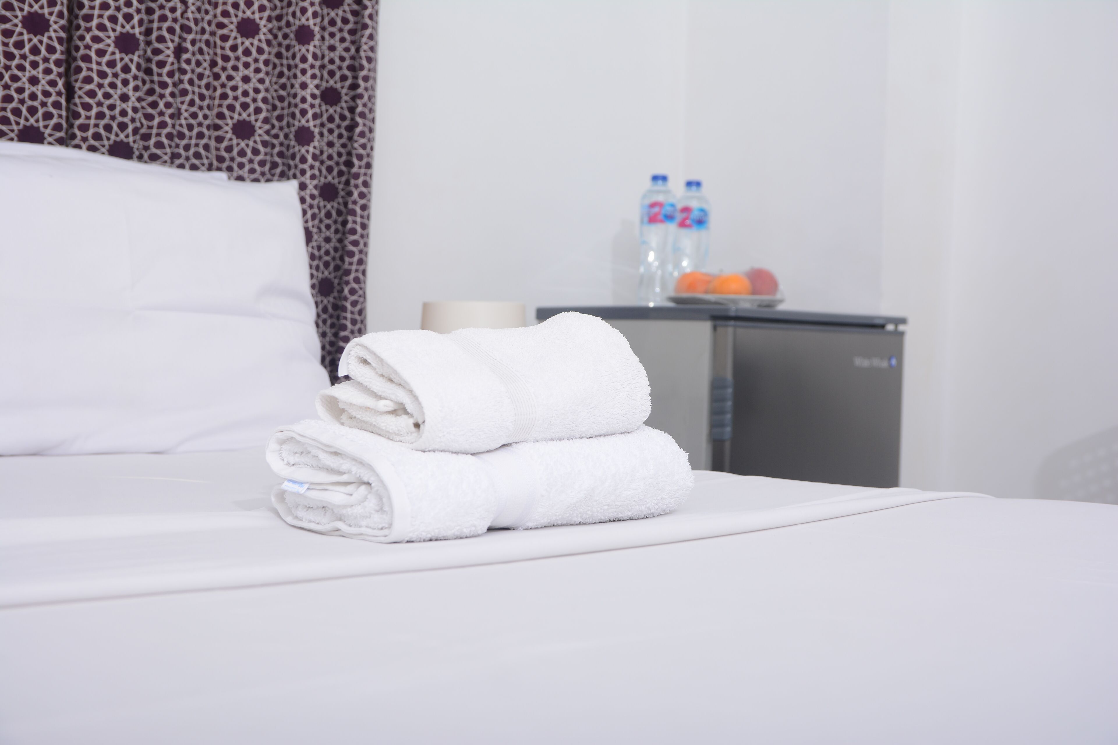 A neatly made bed with folded white towels, a bedside table with two water bottles, and a fruit bowl with oranges.