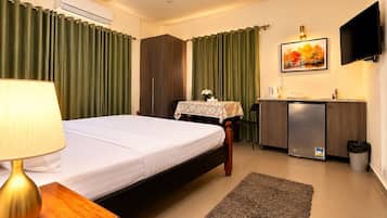 A hotel room with a bed, a desk with a lamp, a small table with a vase, a TV, and a kitchenette area.